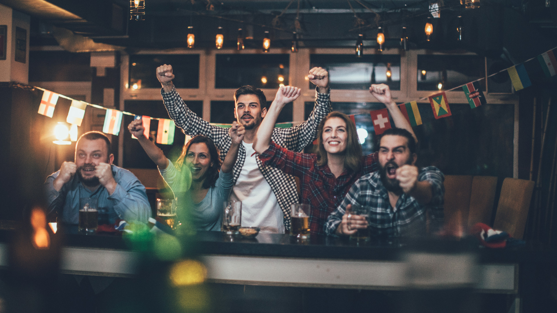 Five people are watching TV at a bar counter and cheering. Fem personer ser på TV vid en bardisk och jublar.